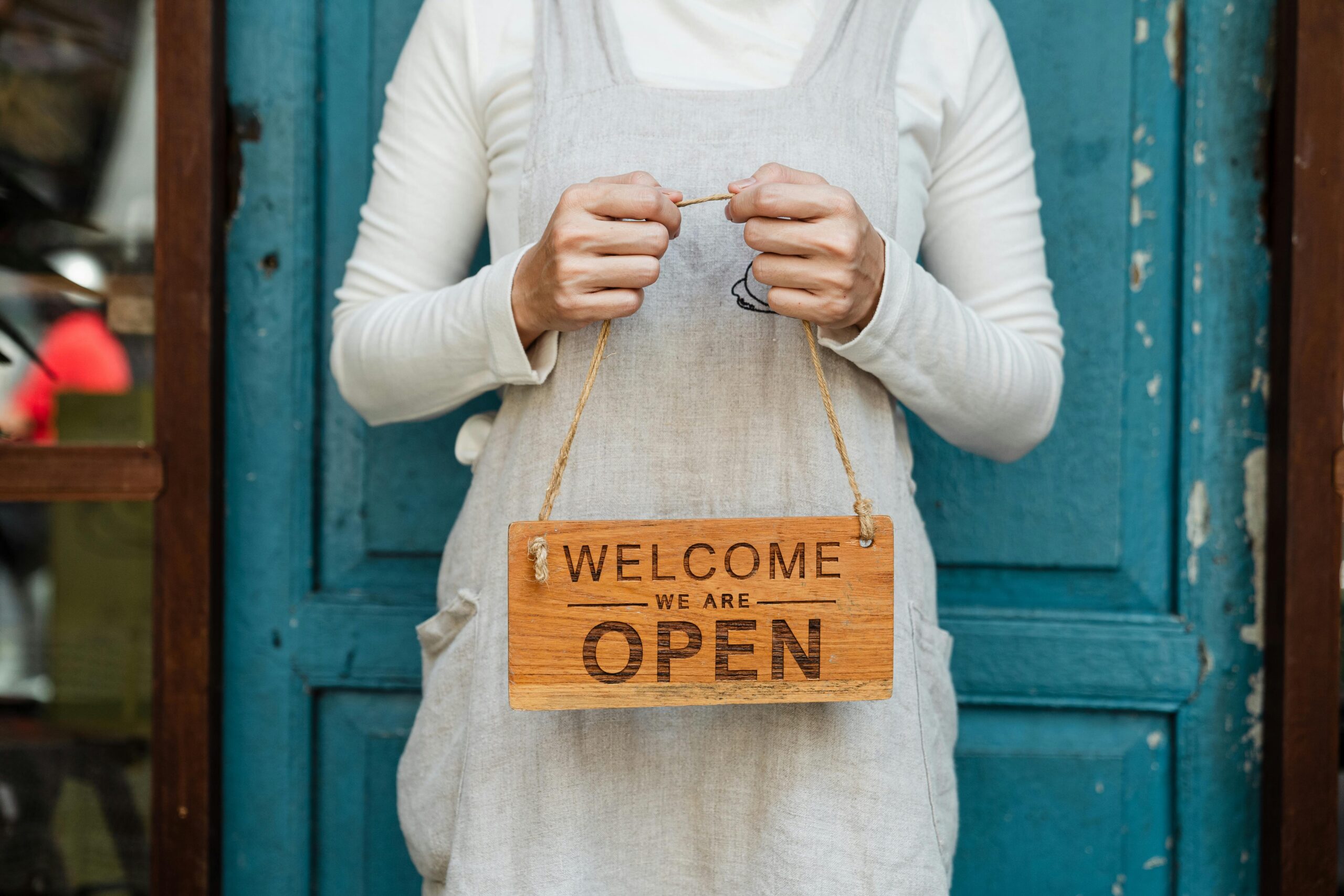 Shop owner holding a 'Welcome, We Are Open' sign in front of a store, representing the need for a retail business loan to support small business operations.